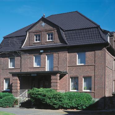 Zweistöckiges Backsteinhaus mit Satteldach, begrüntem Vorgarten und freiem Himmel im Hintergrund.