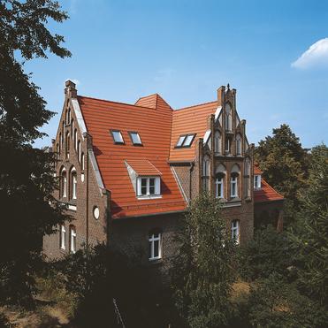 Historisches Gebäude mit rotem Dach und verzierten Fenstern, umgeben von Bäumen und sträucher im Vordergrund.