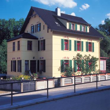 Gelbes Wohnhaus mit roten Fensterrahmen und grünen Fensterläden, umgeben von Bäumen und einem kleinen Garten.