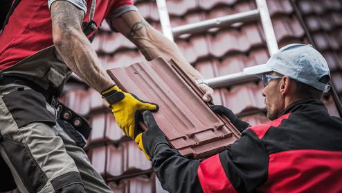 Zwei Handwerker übergeben eine Dachziegel auf einem Steildach, während eine Leiter im Hintergrund sichtbar ist.