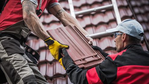 Zwei Handwerker übergeben eine Dachziegel auf einem Steildach, während eine Leiter im Hintergrund sichtbar ist.