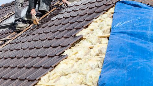 Handwerker verlegt Dachziegel auf einem Dach, während isolierendes Material und eine blaue Plane sichtbar sind.