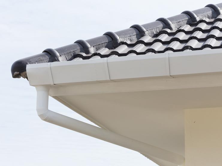 Detailaufnahme einer Dachkante mit schwarzer Ziegeldeckung und weißer Regenrinne unter einem grauen Himmel.