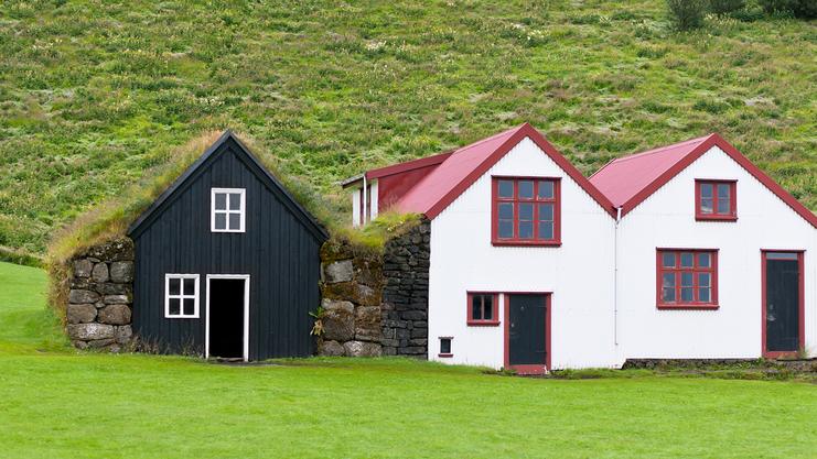 Zwei traditionelle isländische Häuser mit bunten Dächern stehen vor einer grünen Hügelkulisse.